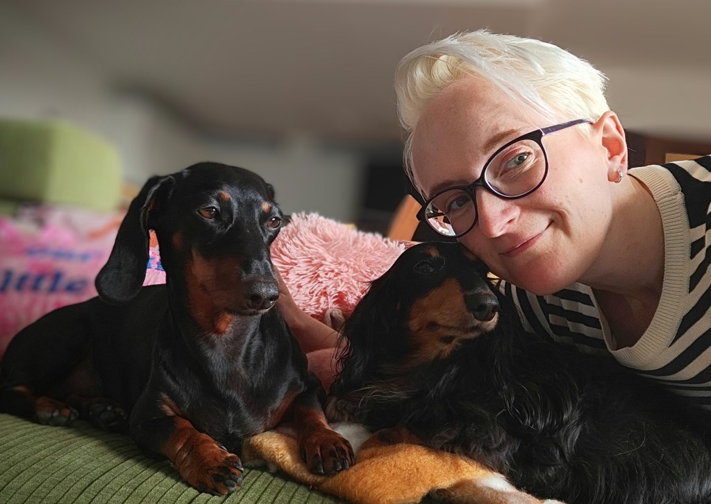 A person with short blonde hair and glasses sits on a couch with two dachshund dogs, one black and tan and the other black. The background features a colorful blanket and a plush green sofa.
