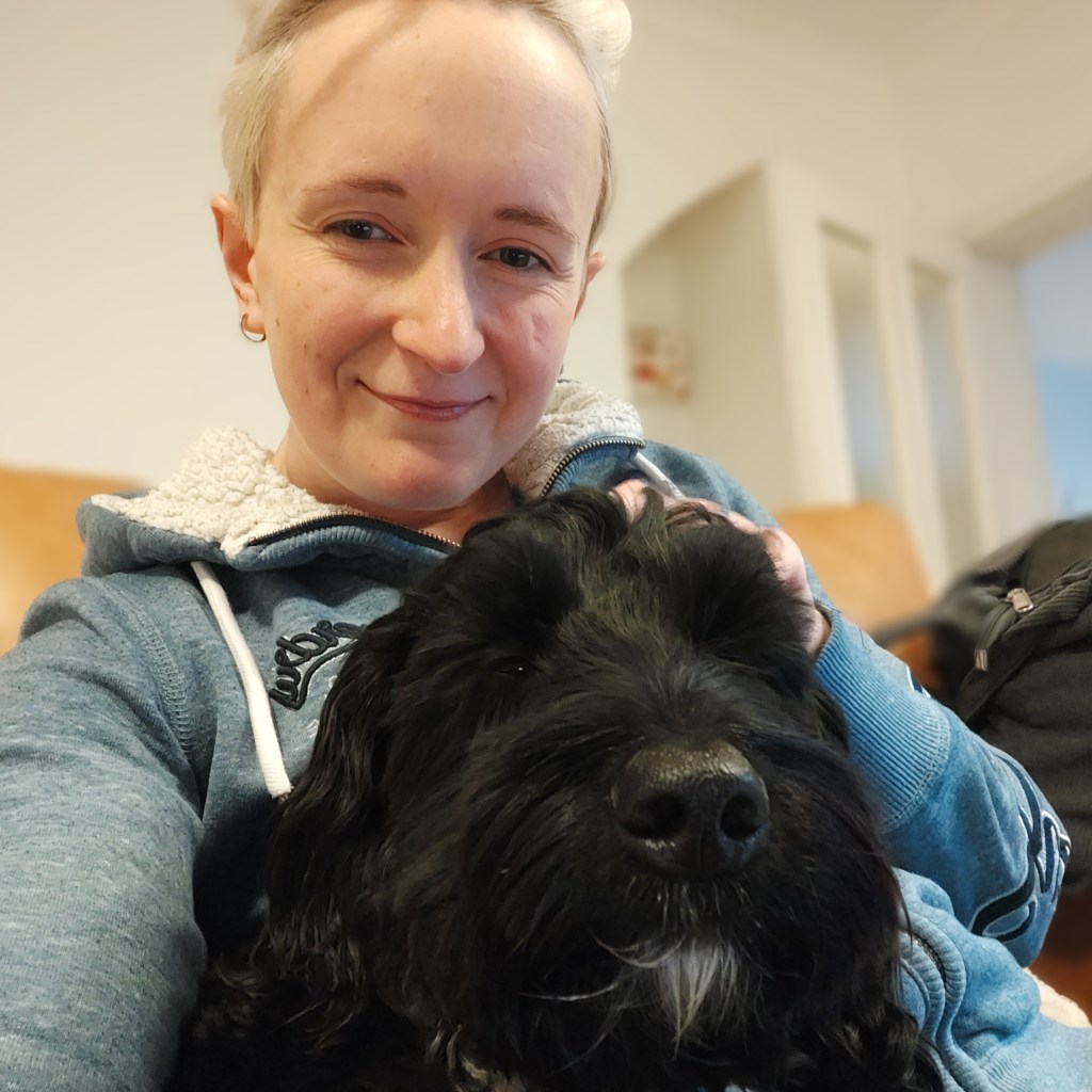 A person with short light hair smiling while sitting on a sofa, holding a black dog close to them.