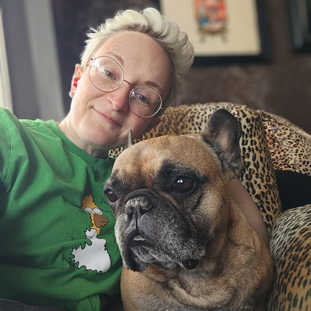 A person with short hair wearing glasses and a green shirt poses for a selfie with a brown French Bulldog, both looking at the camera while sitting on a leopard print chair.