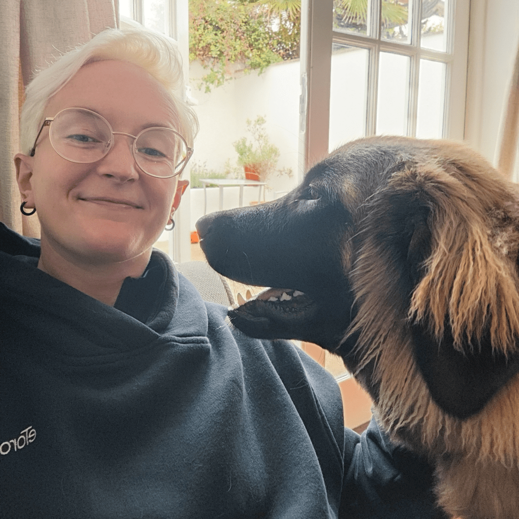 A person with short blonde hair and glasses smiling while interacting with a dog indoors, showing a warm and affectionate moment.
