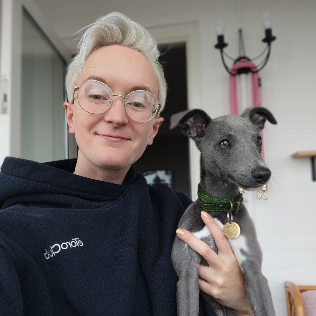 A person with short platinum blonde hair and glasses is holding a small grey dog with a green collar. They are smiling at the camera, sitting indoors.