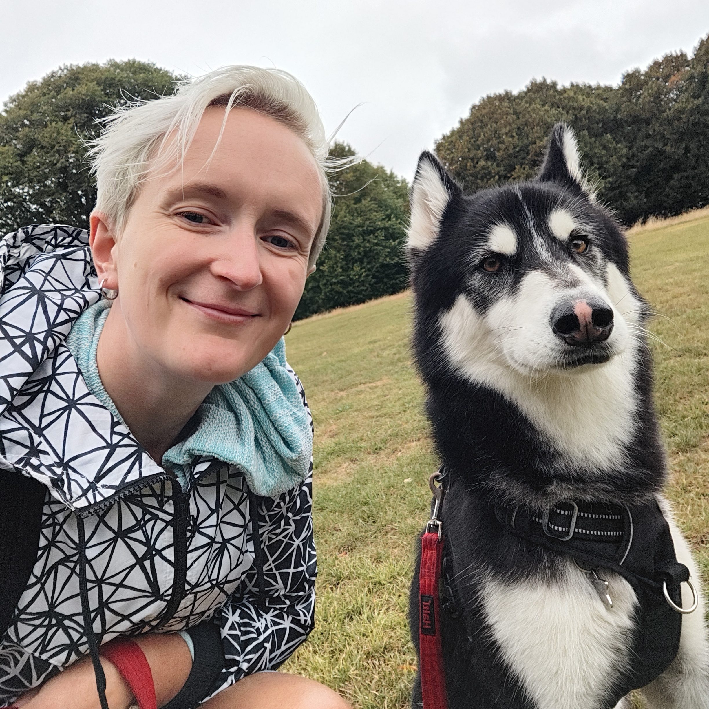 A person with short hair and a patterned jacket smiling next to a black and white dog in a grassy field.