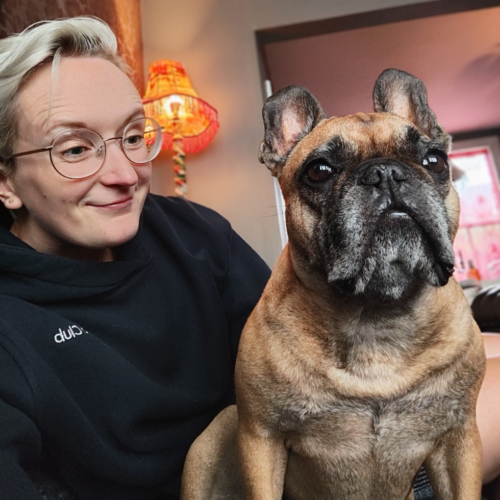 A person with short light hair wearing glasses is sitting next to a brown French Bulldog, both looking content in a cozy living space.